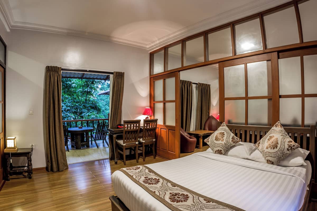 Habitación de hotel con una cama grande, un escritorio con silla, una mesita con otra silla, y vista a zonas verdes por una ventana.