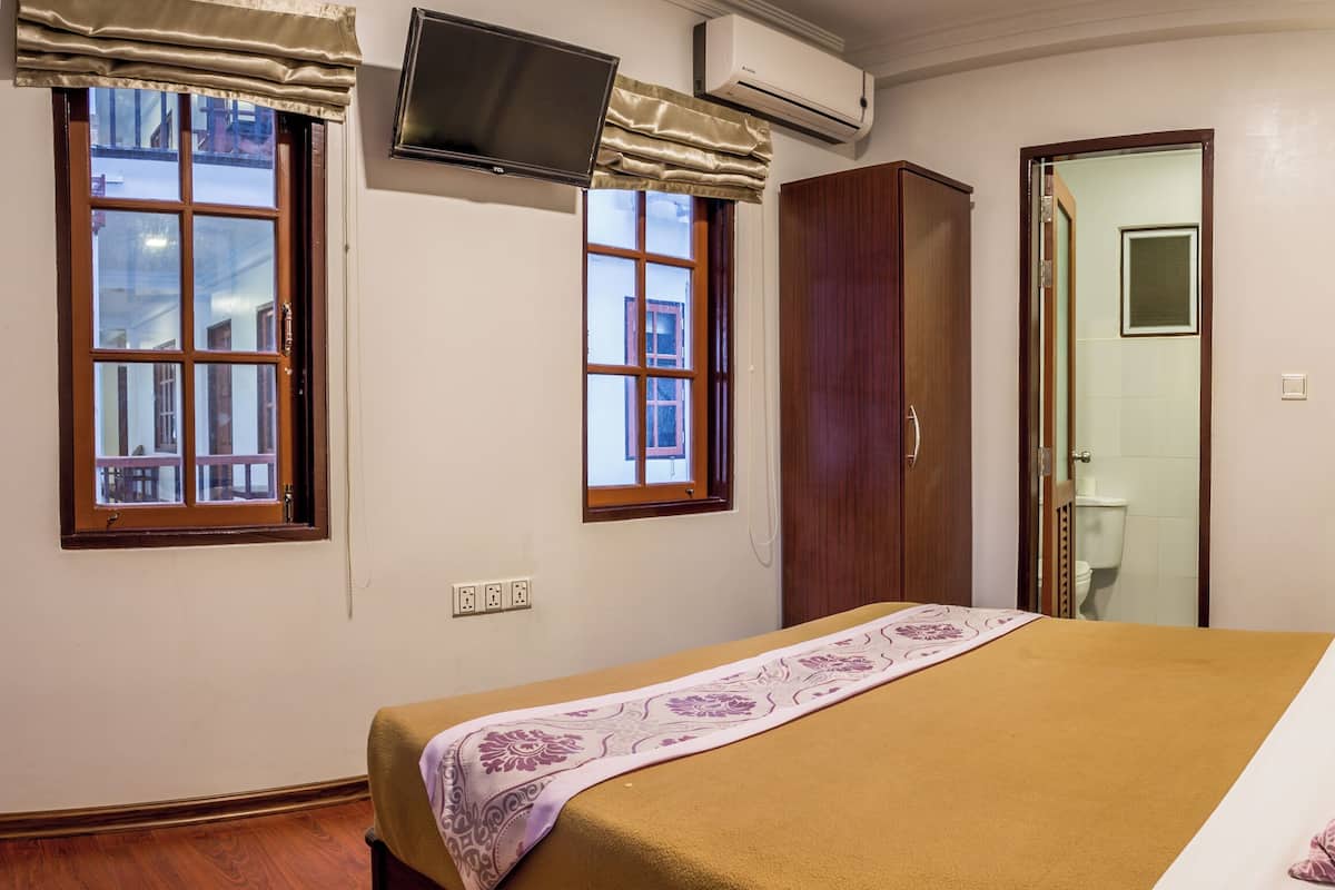 Habitación de hotel con cama, televisión montada en la pared, unidad de aire acondicionado, ventana con cortinas y puerta que da a un baño.