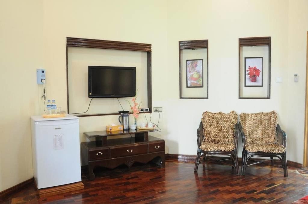 A room with a flat-screen TV mounted on the wall, a small refrigerator, a wooden cabinet with drawers, two wicker chairs, and framed pictures on the wall.