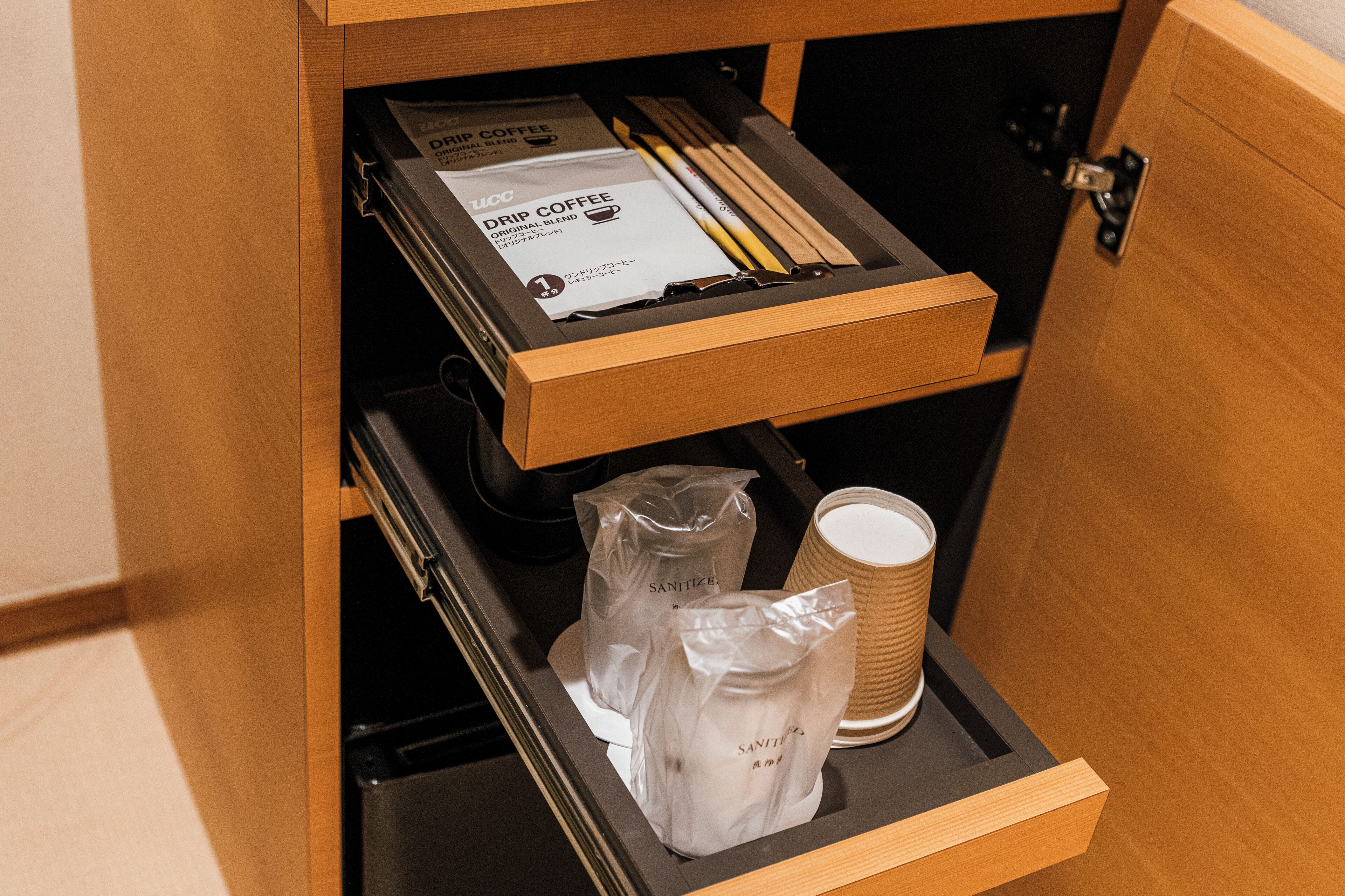 A drawer with a drip coffee box, two plastic bags labeled 'Sanitizer', and a stack of paper cups.