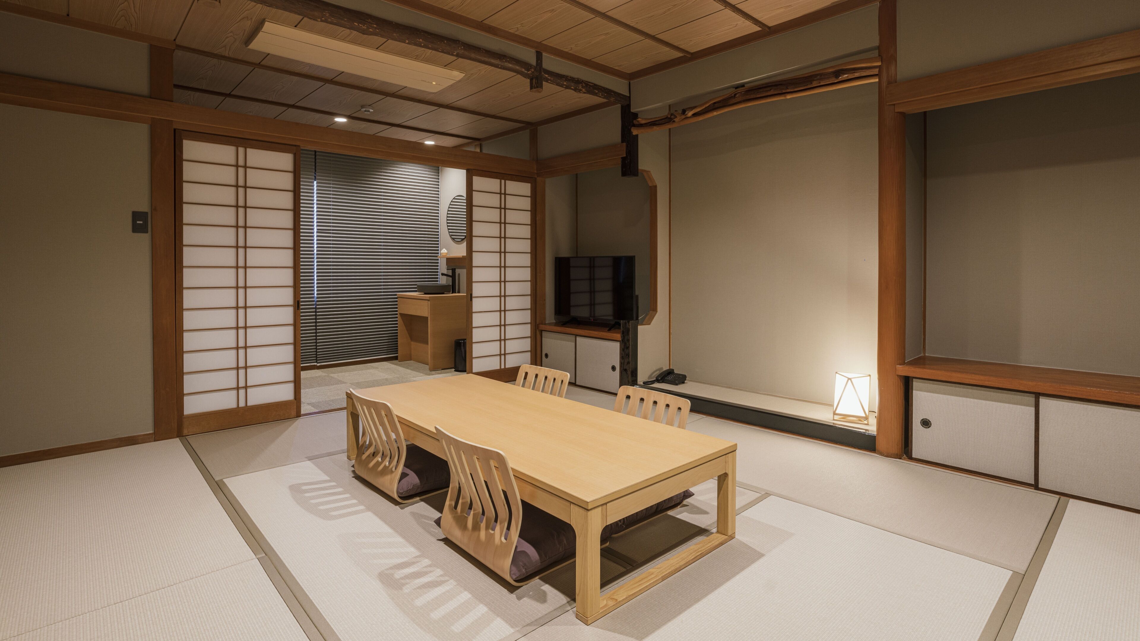 A traditional Japanese-style room with tatami flooring, a wooden table, chairs, a television, and sliding doors.