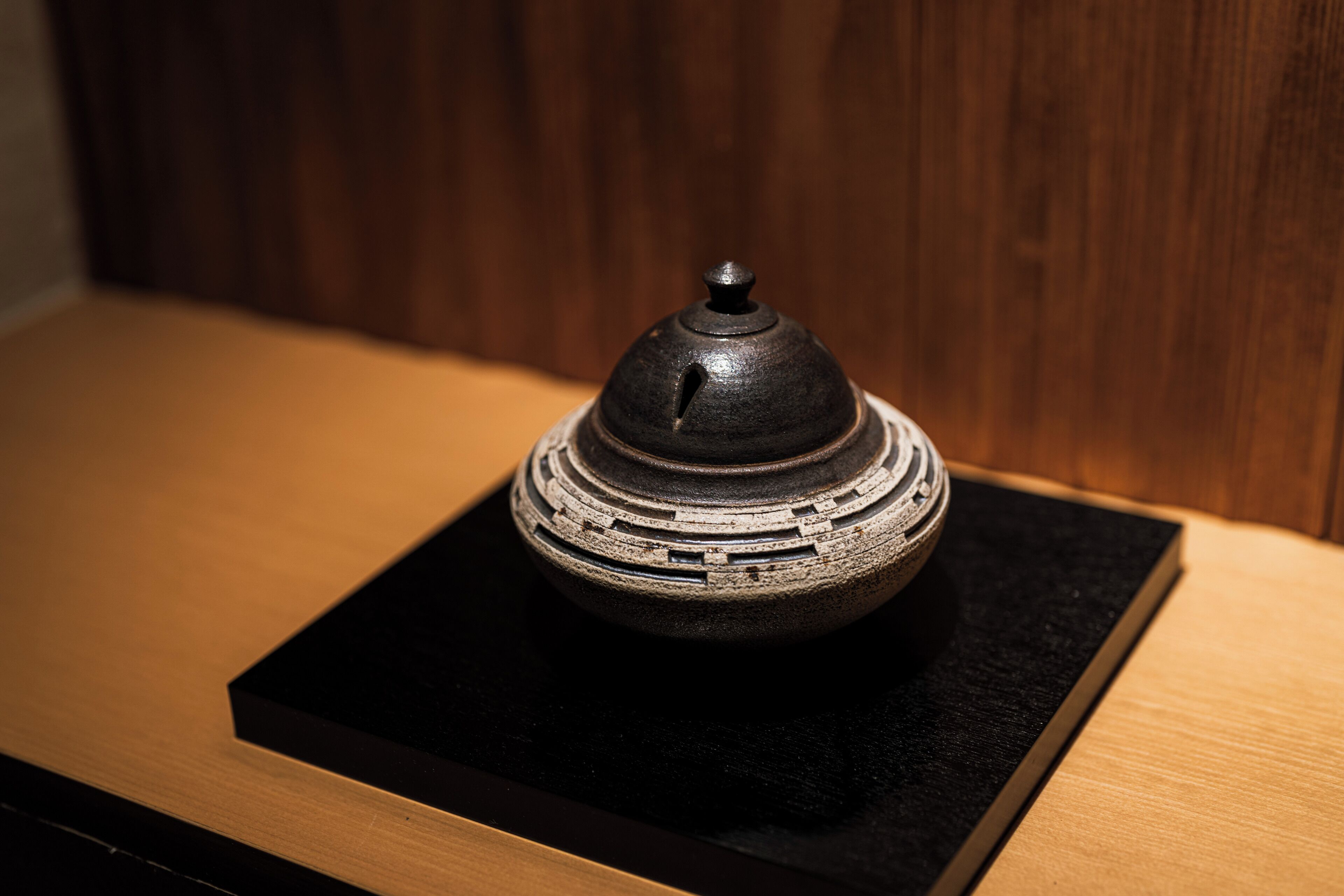 A small, dark-colored ceramic object with a lid, placed on a black square base, against a wooden background.