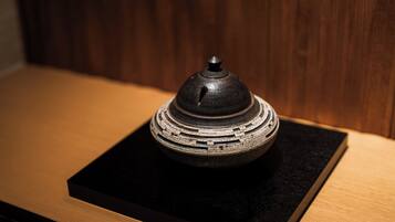 A small, dark-colored ceramic object with a lid, placed on a black square base, against a wooden background.