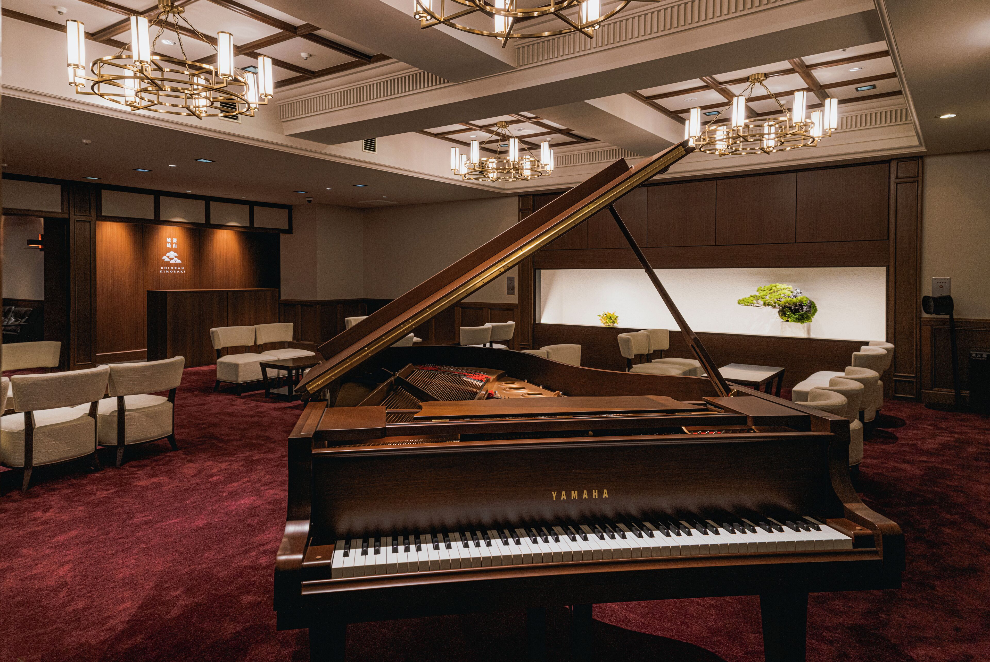 A grand piano in a hotel lobby with a red carpet, seating area, and a large screen displaying a floral arrangement.