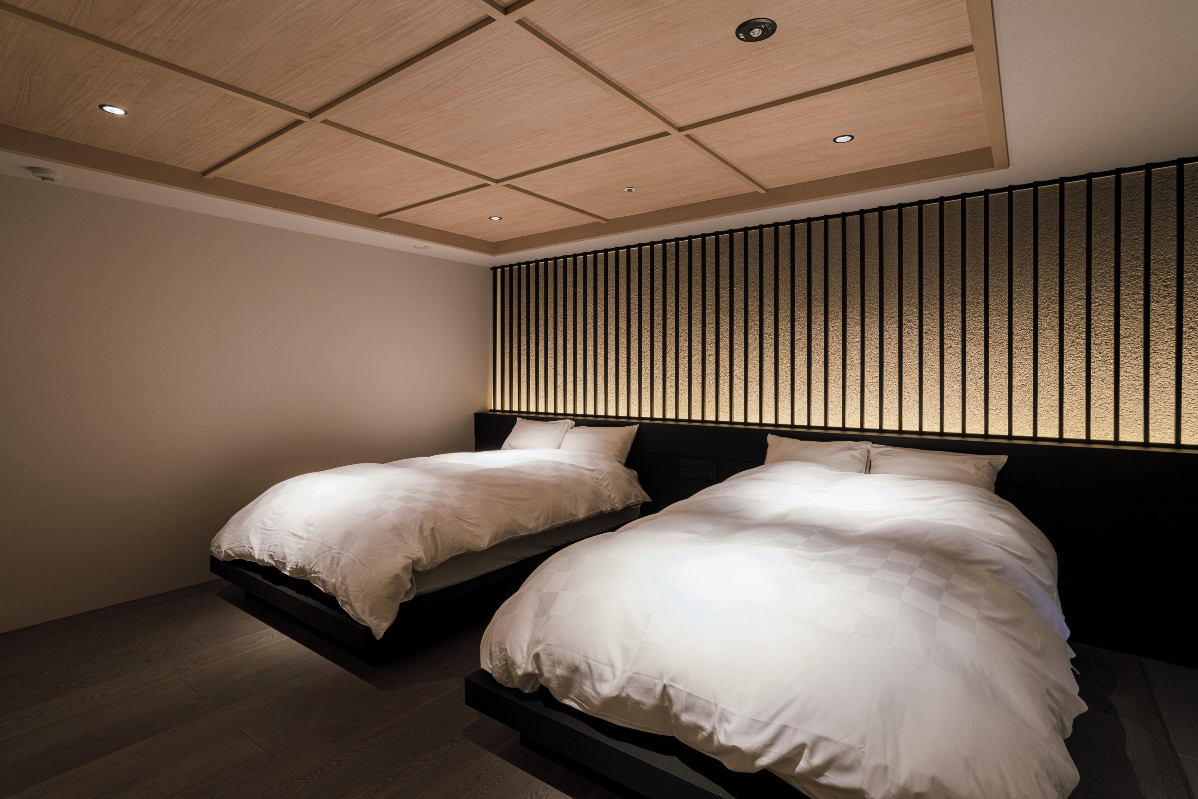 A bedroom with two beds, wooden flooring, a slatted headboard, and recessed ceiling lighting.