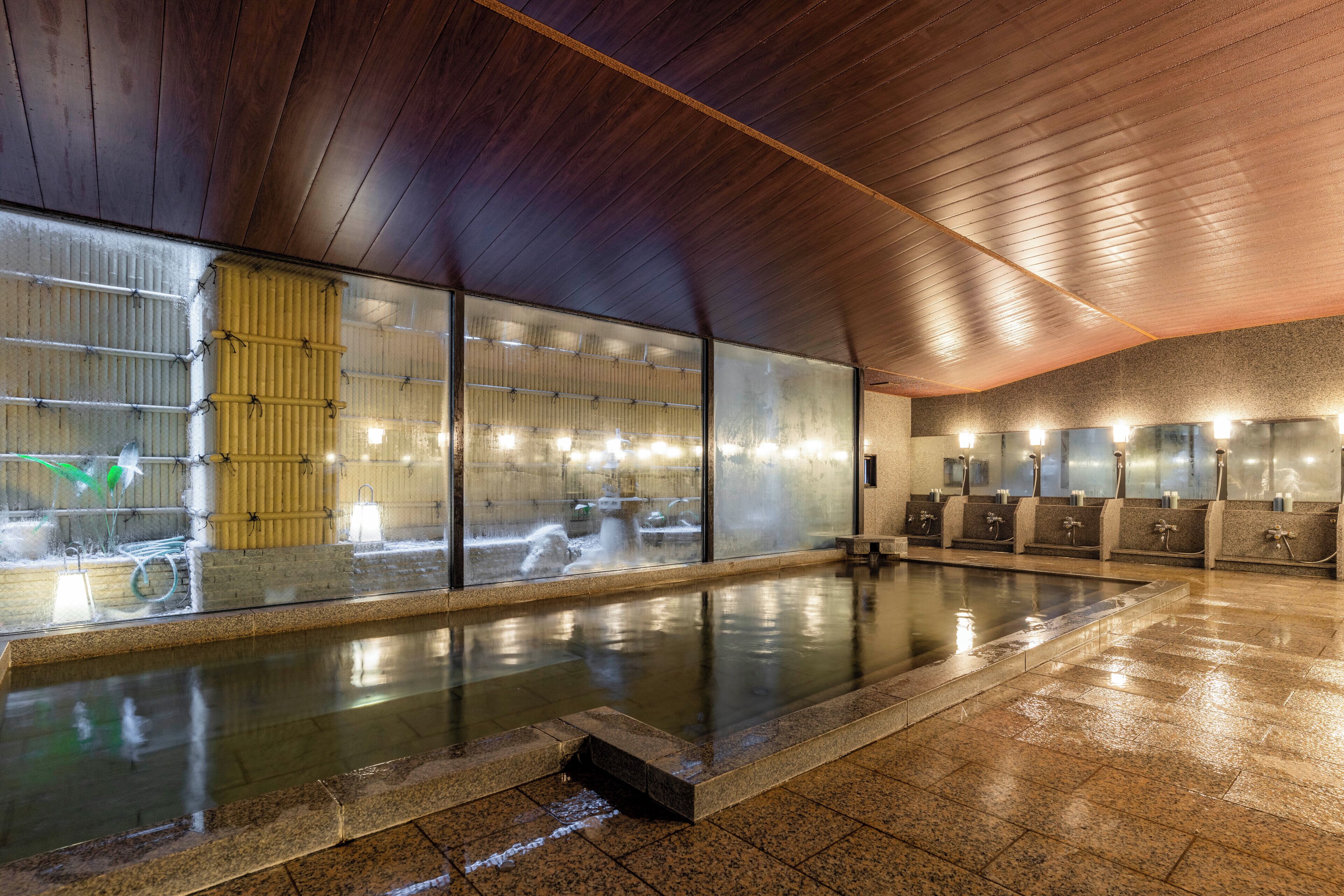 A large indoor swimming pool with a wooden ceiling and tiled floor, surrounded by glass walls and multiple shower stations.