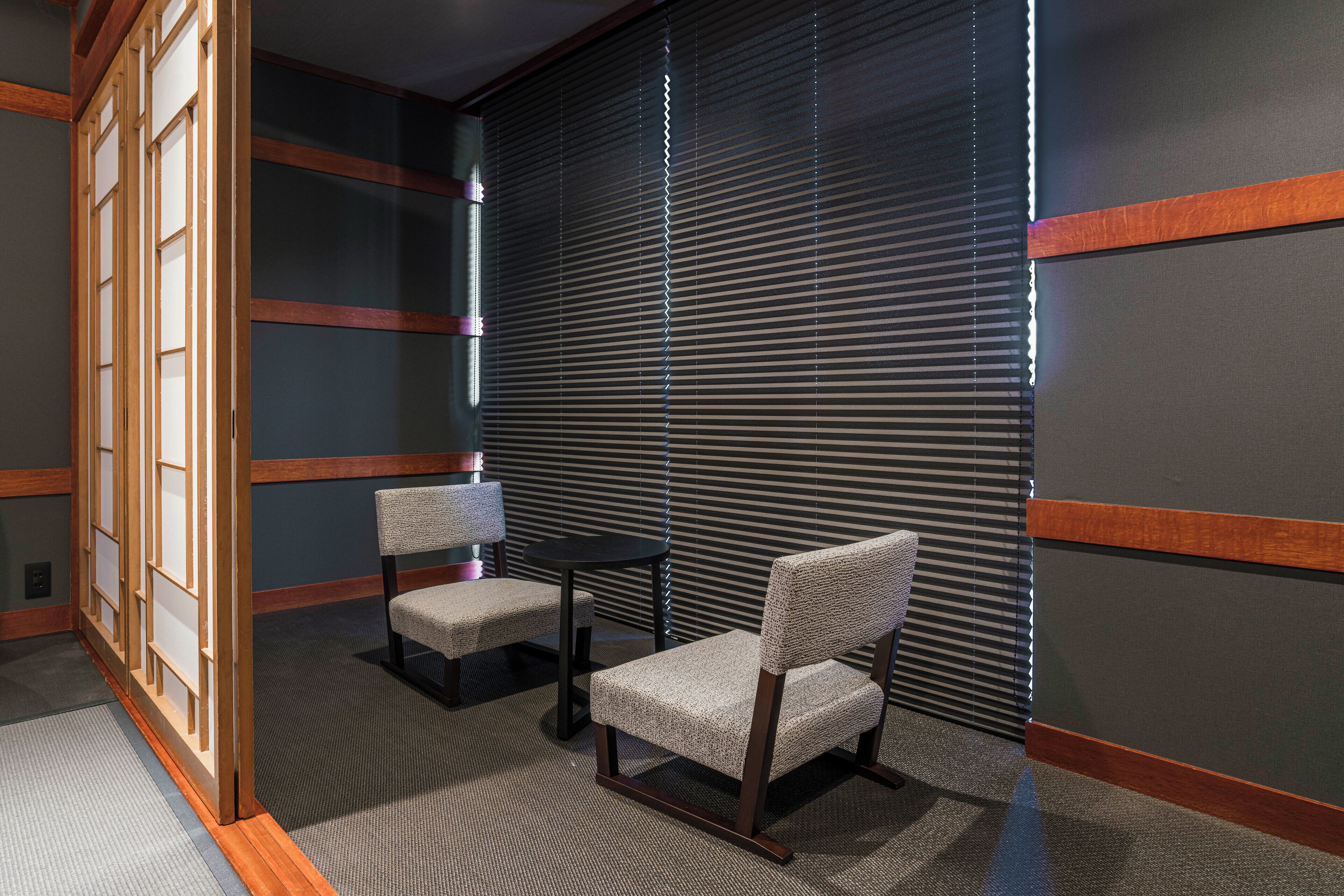 Una habitación con dos sillas, una mesita y una puerta corredera, y una pared oscura con líneas horizontales.