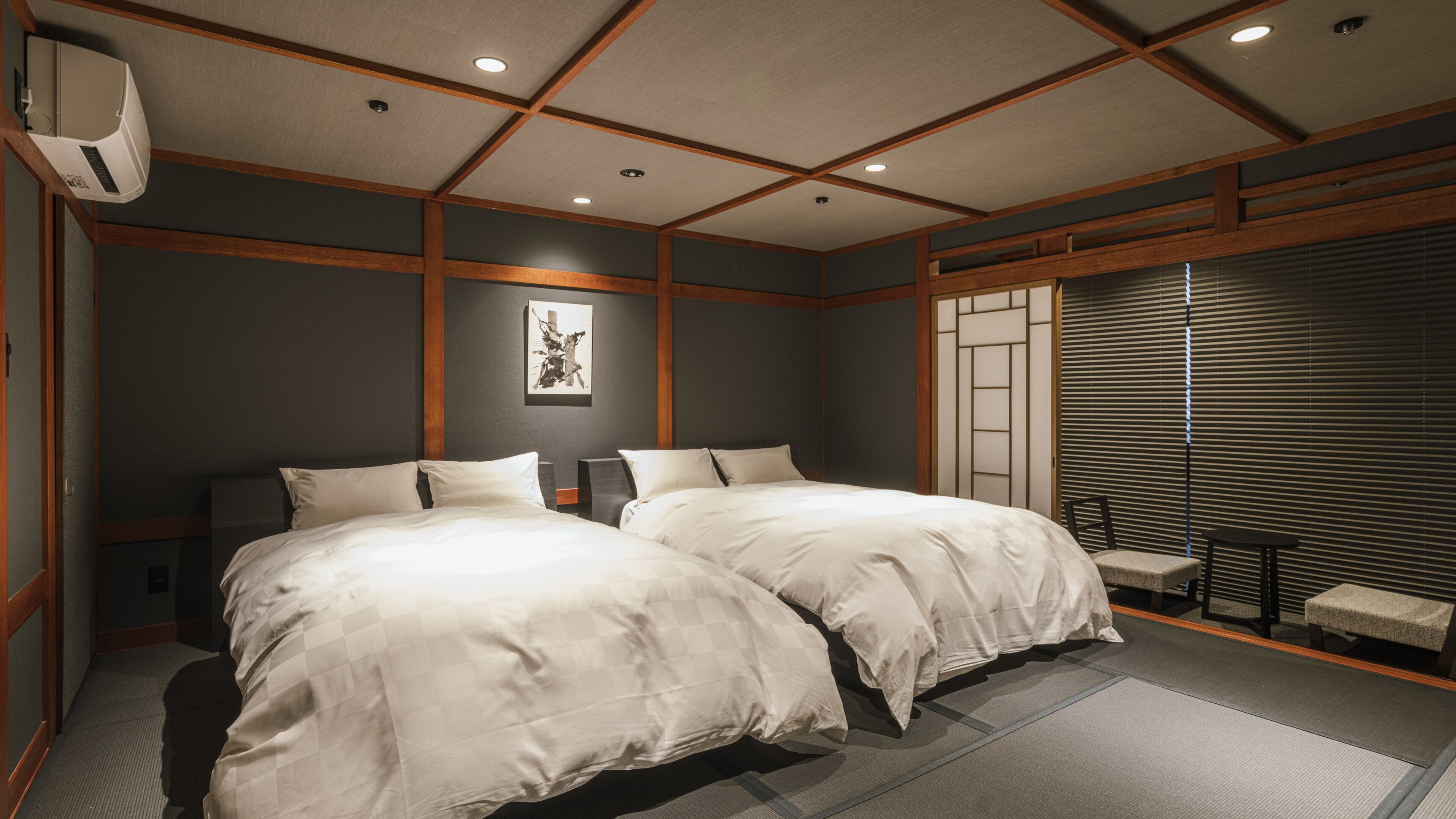 A hotel room with two beds, a small table, a chair, a wall-mounted air conditioner, and a sliding door.