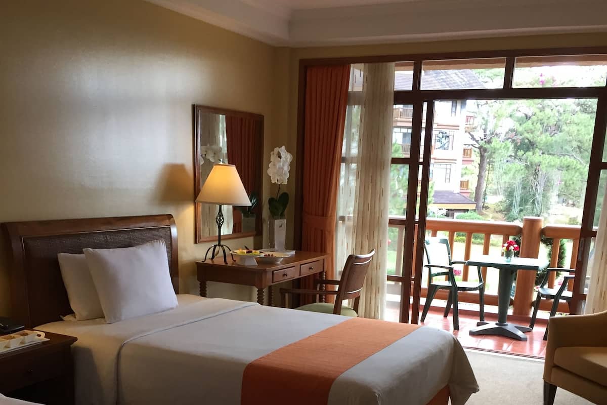 Quarto de hotel com duas camas, uma escrivaninha, uma cadeira e varanda com mesa e cadeiras.