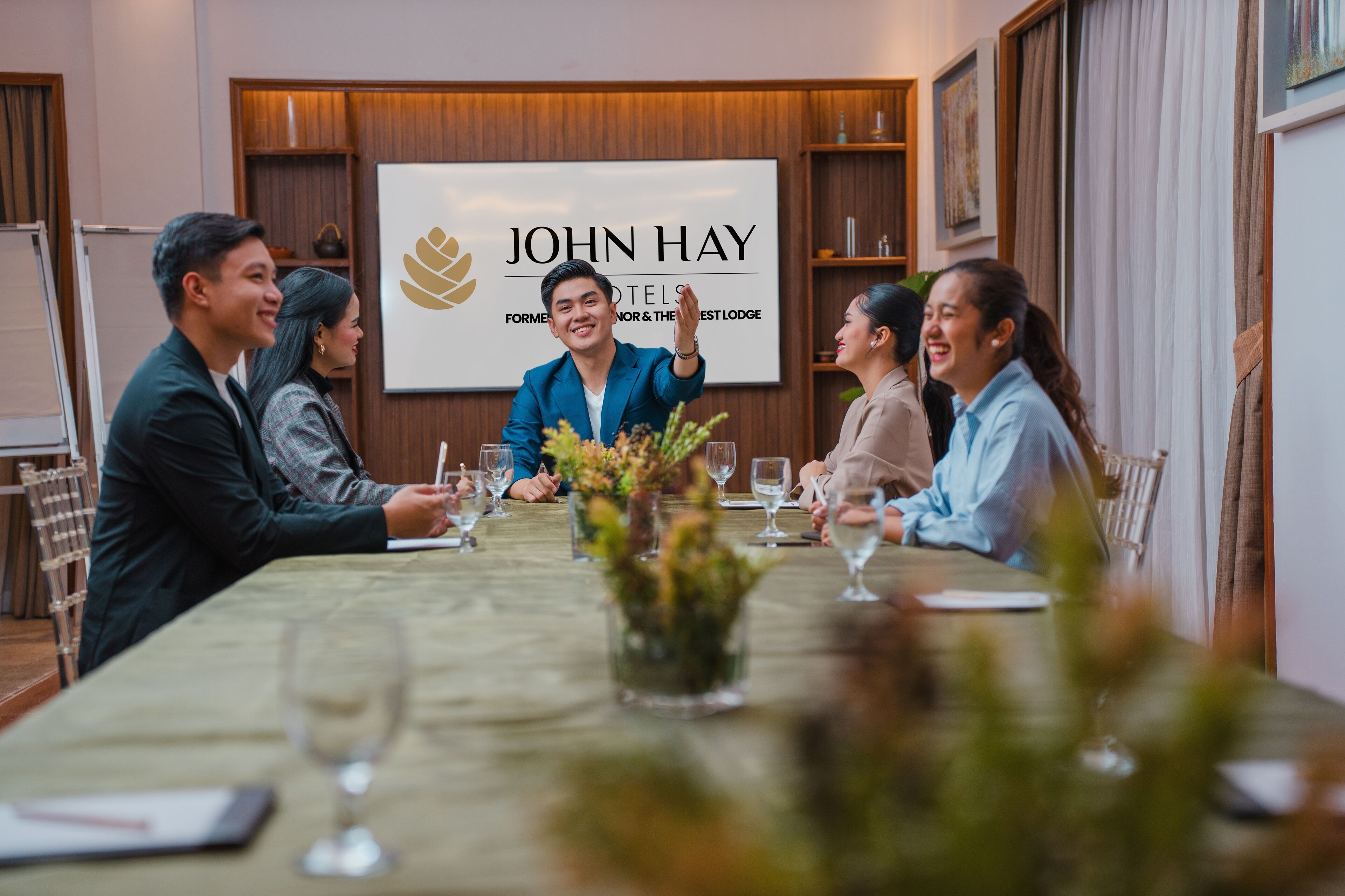 A conference room with a long table, chairs, and a large screen displaying the John Hay Hotels logo.