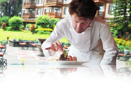 A chef preparing food outdoors with a view of a building and greenery in the background.