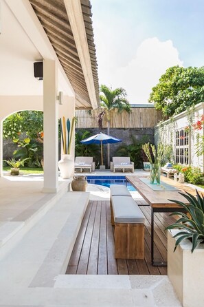 A covered outdoor area with a pool, wooden benches, lounge chairs, and potted plants. - Allure Villas (Seminyak)