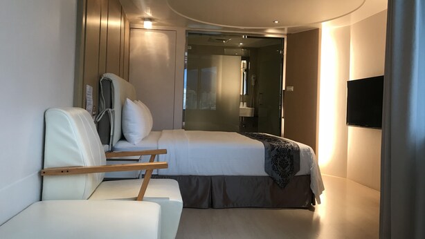 A modern hotel room with a bed, a seating area with a chair and a small table, a TV mounted on the wall, and a shower area visible through a glass door.