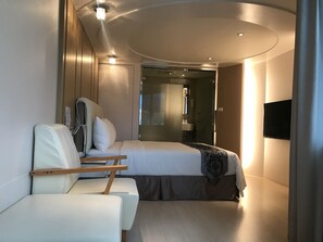 Quarto de hotel moderno com uma cama, área de estar com uma cadeira e uma pequena mesa, TV fixada na parede e área de chuveiro visível através de uma porta de vidro.