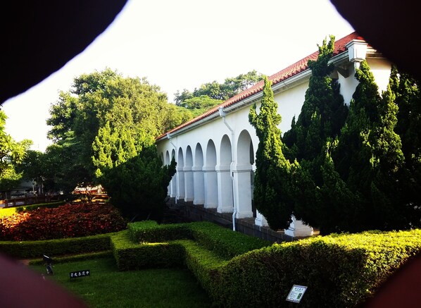 Um edifício com uma fileira de arcos e um telhado de telhas vermelhas, rodeado por sebes e árvores bem cuidadas.