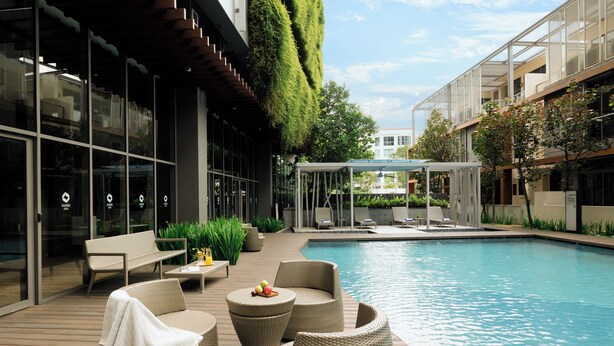 Un área de piscina moderna al aire libre con mobiliario de mimbre, una zona de estar cubierta y un edificio de cristal con pared verde.