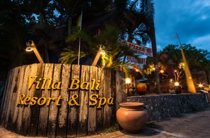 Un letrero de madera con la inscripción 'Villa Bali Resort & Spa', rodeado de palmeras e iluminación exterior.