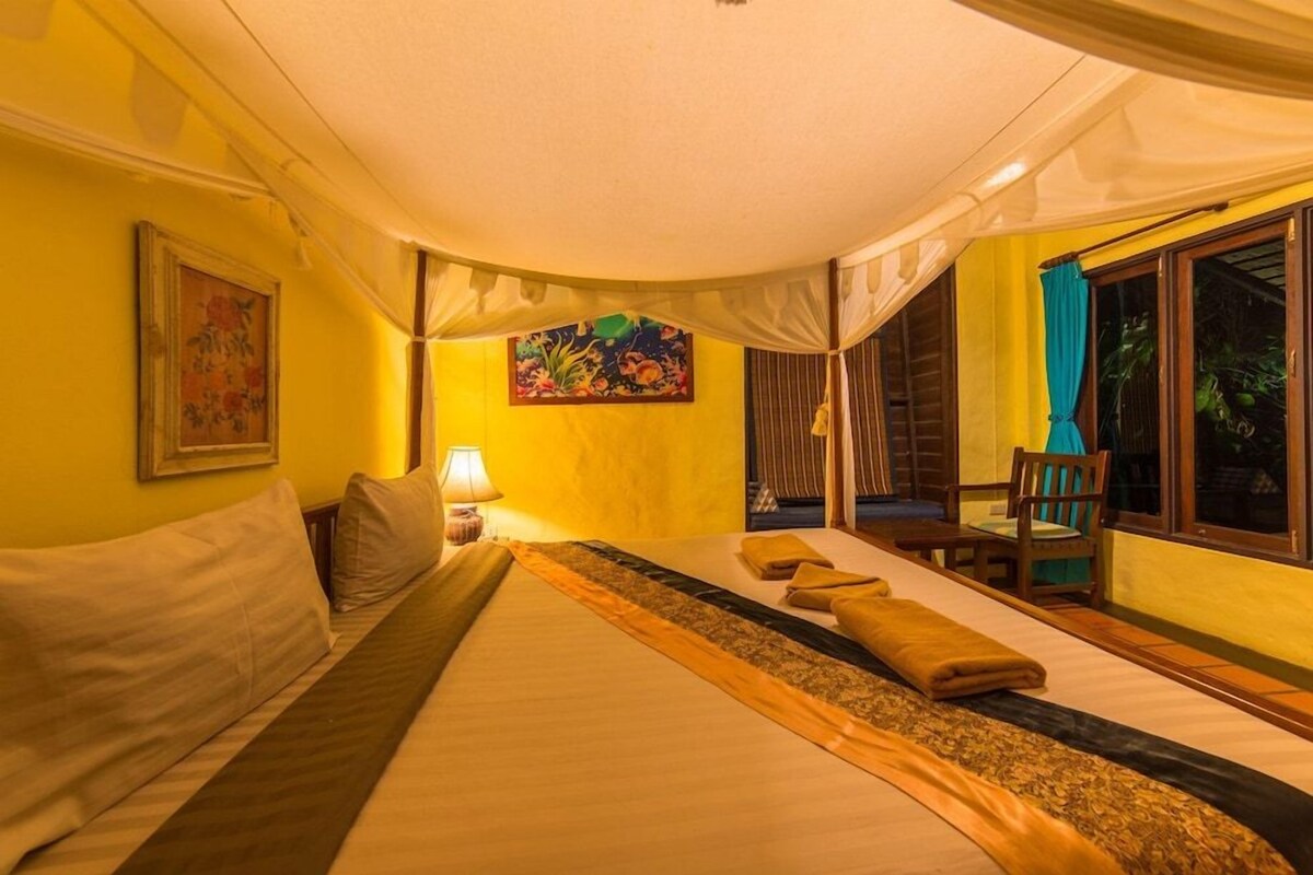 A room with a bed, a painting, a lamp, and a view of the outdoors.