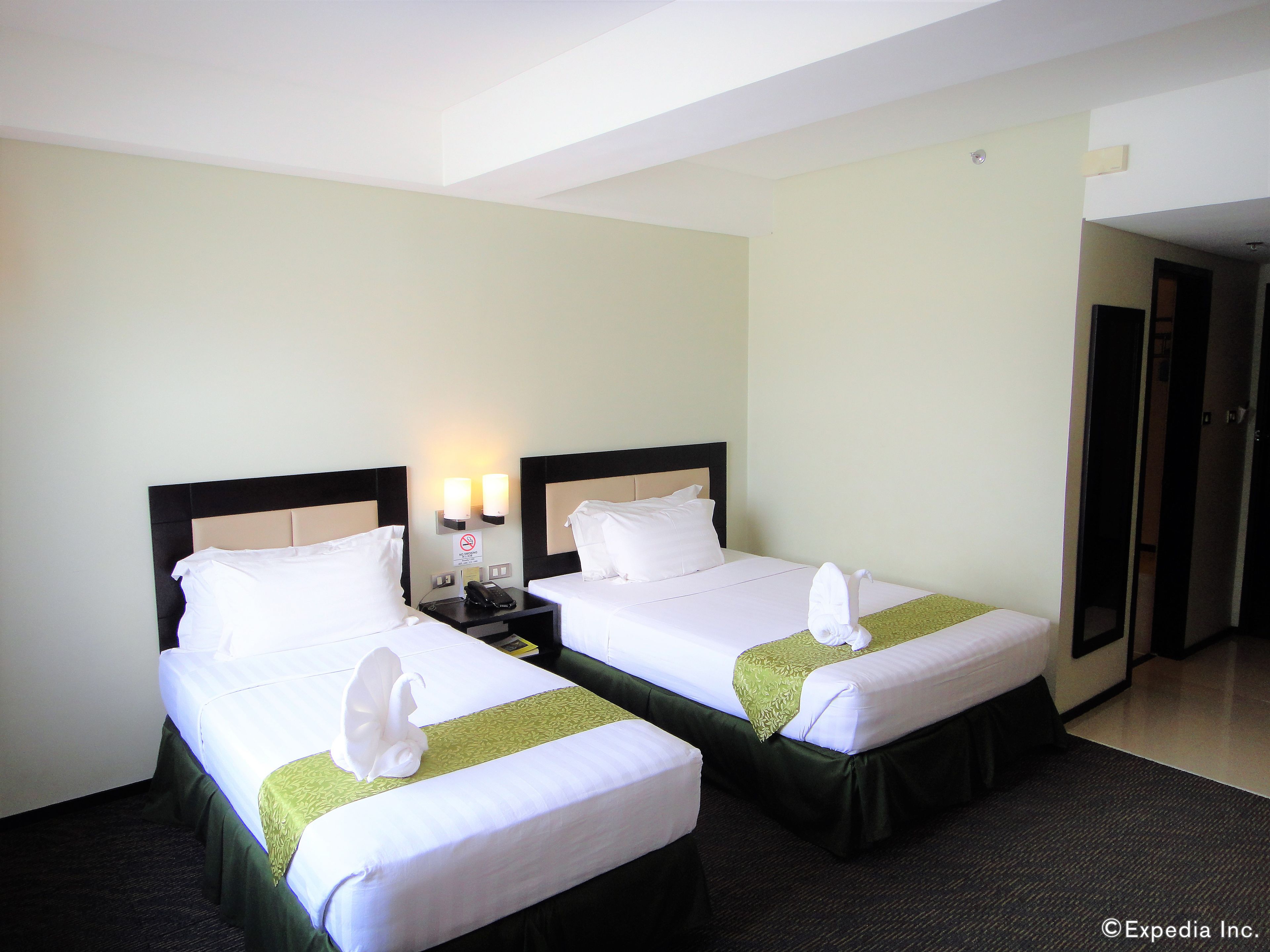 A hotel room with two beds, each with white linens and a green bed skirt. There are two bedside tables with lamps and a no-smoking sign.