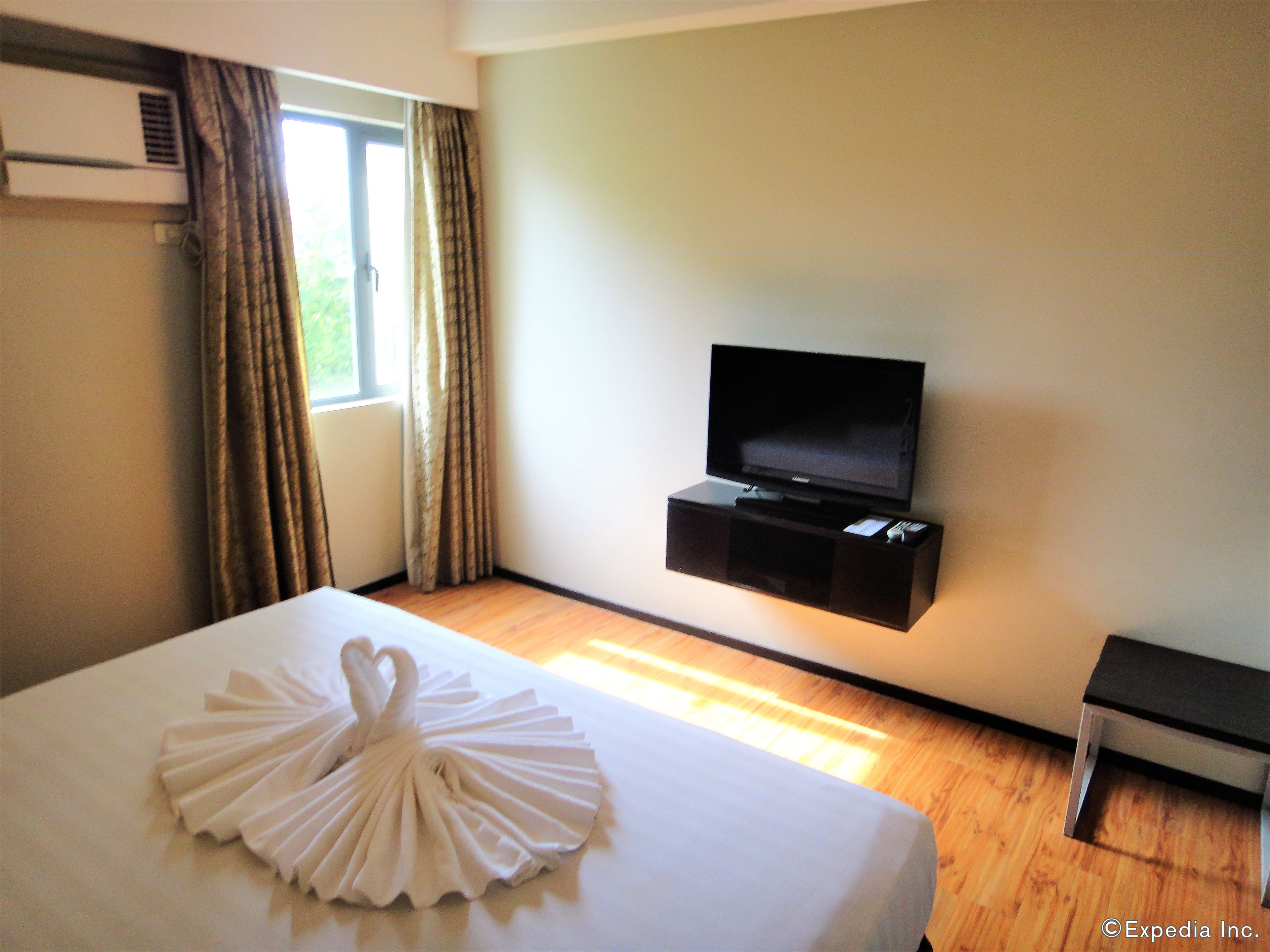 A hotel room with a bed, a flat-screen TV mounted on the wall, a window with curtains, and a wooden floor.