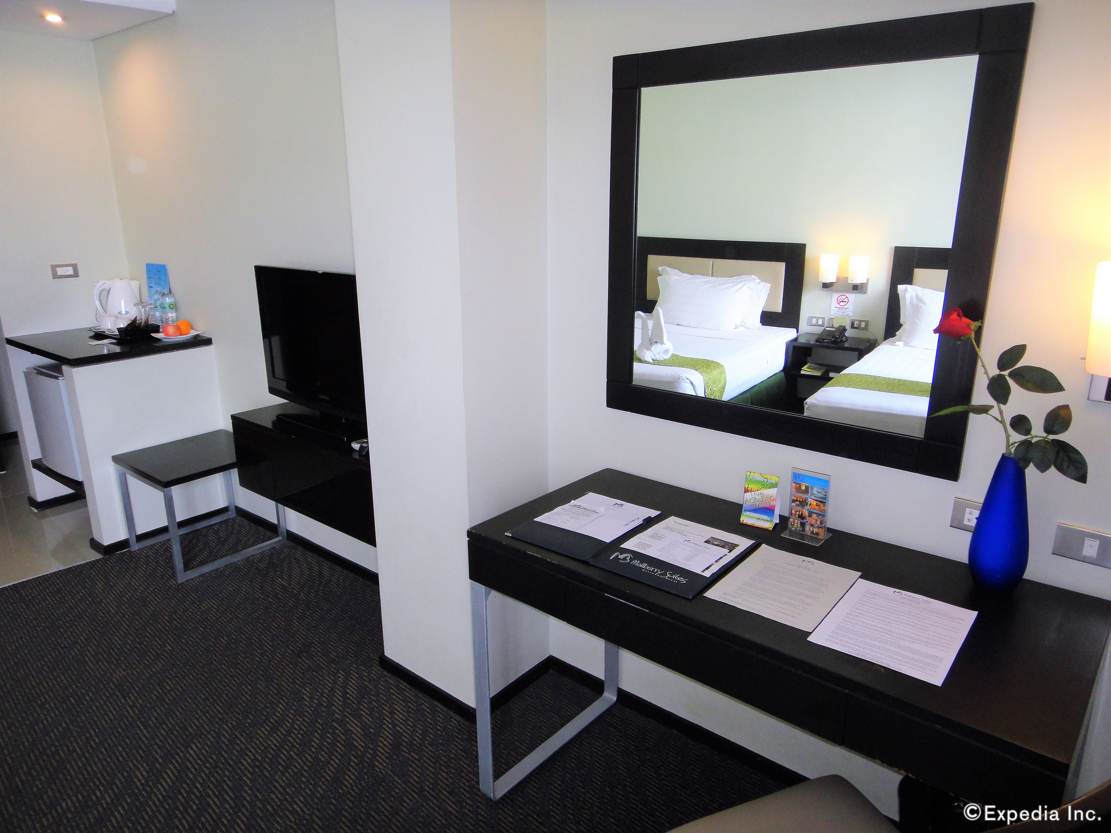 A hotel room with a desk, a mirror, a vase with a flower, and a bed with a green bedspread.