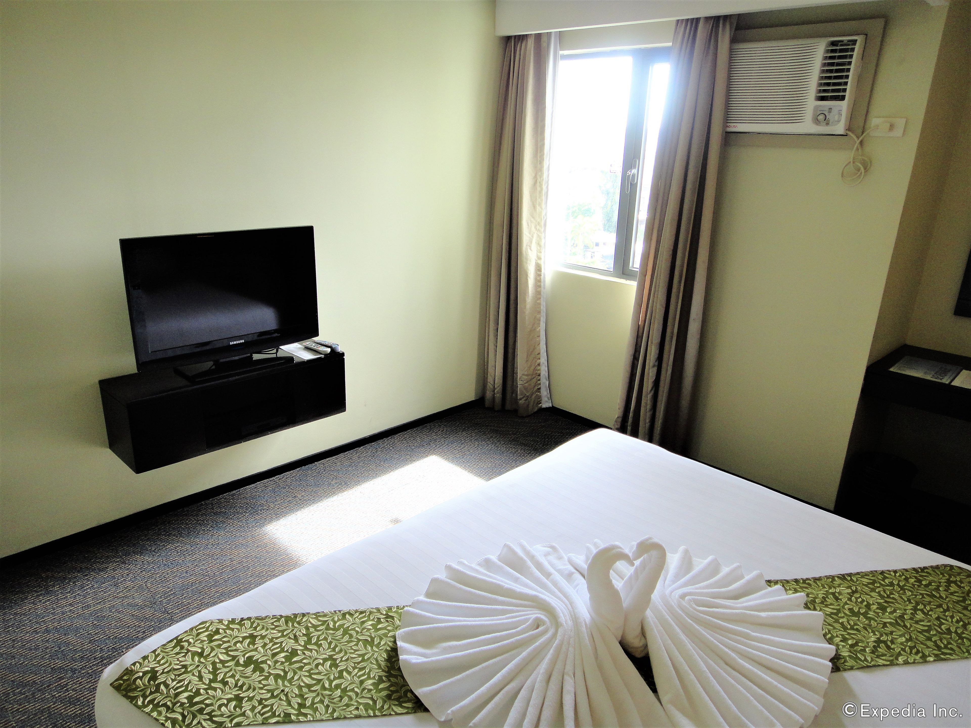 A hotel room with a bed, a television on a stand, a window with curtains, and an air conditioning unit.