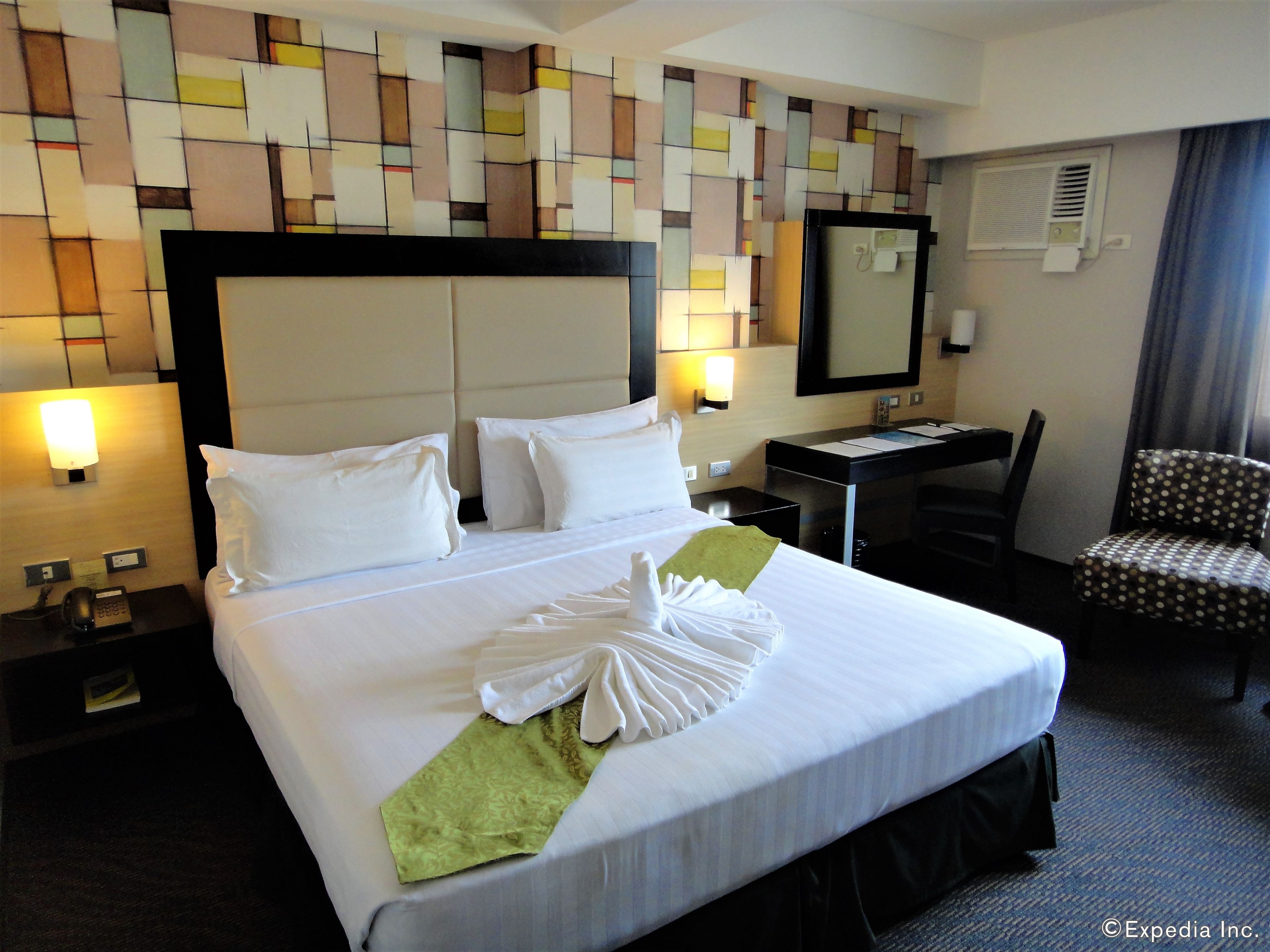 A hotel room with a neatly made bed, a desk with a chair, a mirror, and a wall with a geometric pattern.