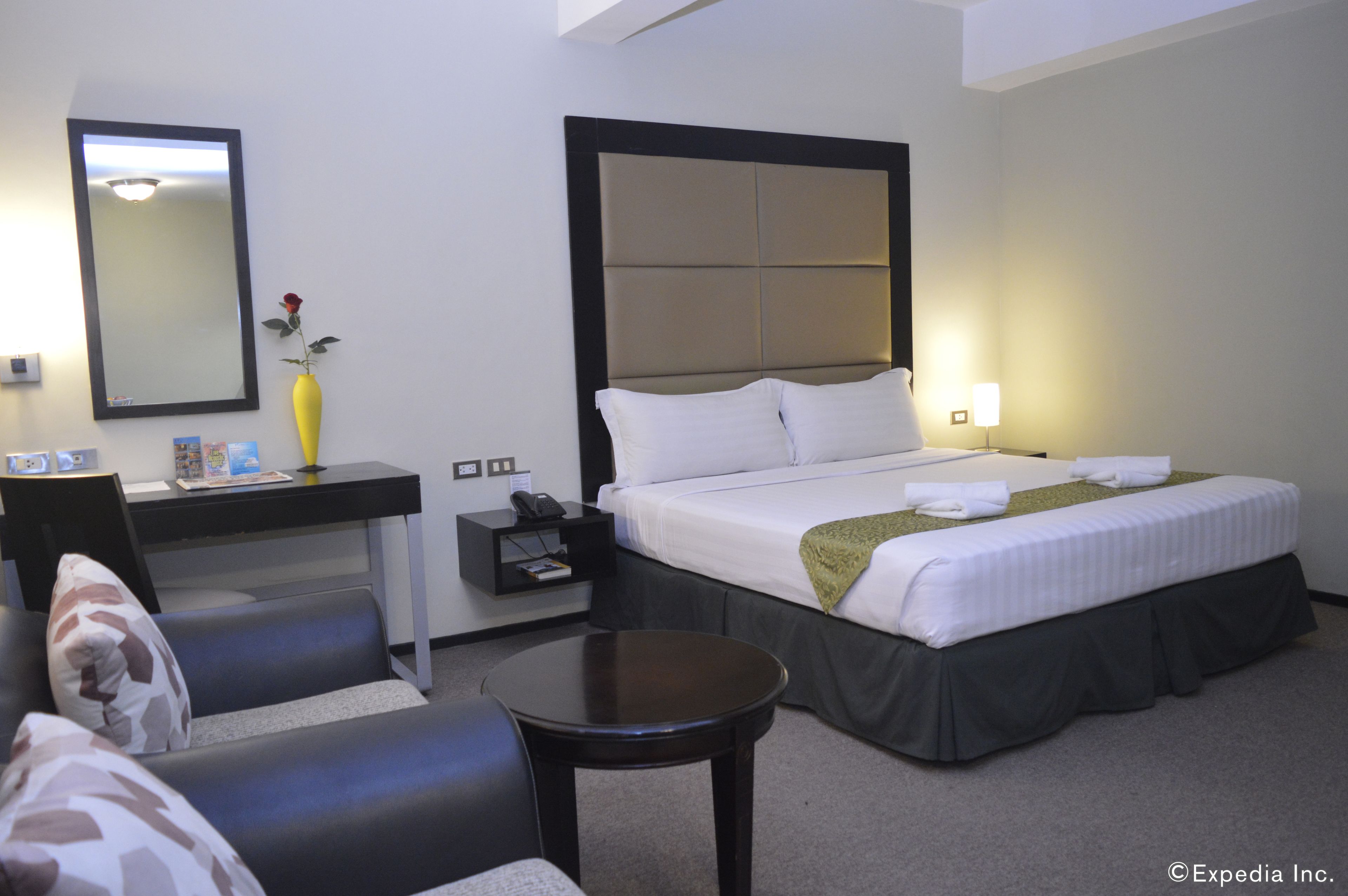 A hotel room with a bed, a desk, a chair, a small table, a nightstand, a mirror, and a vase with a flower.