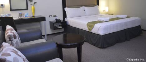 A hotel room with a bed, a desk, a chair, a small table, a nightstand, a mirror, and a vase with a flower.