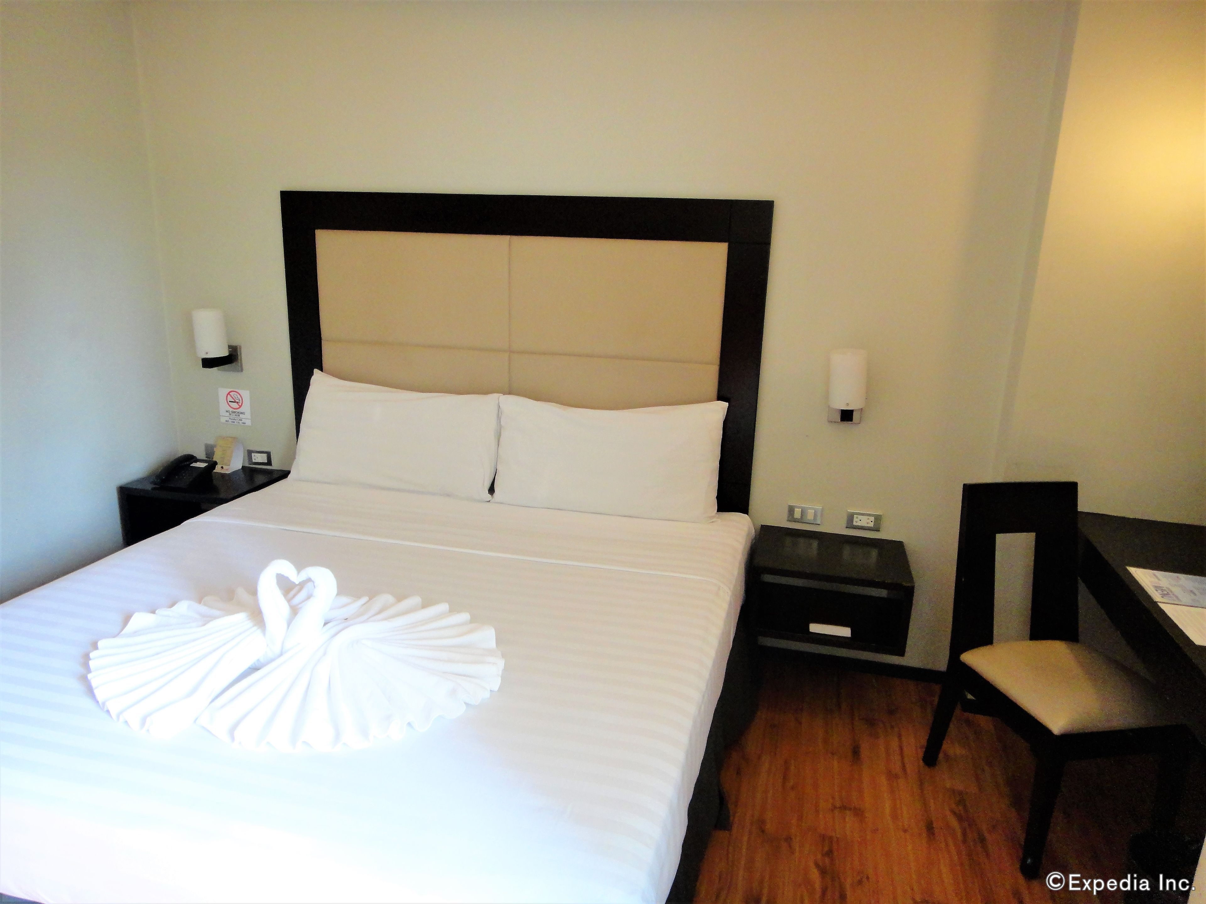 A neatly made bed with a swan-shaped towel, a wooden nightstand with a phone and a lamp, and a chair with a cushion.