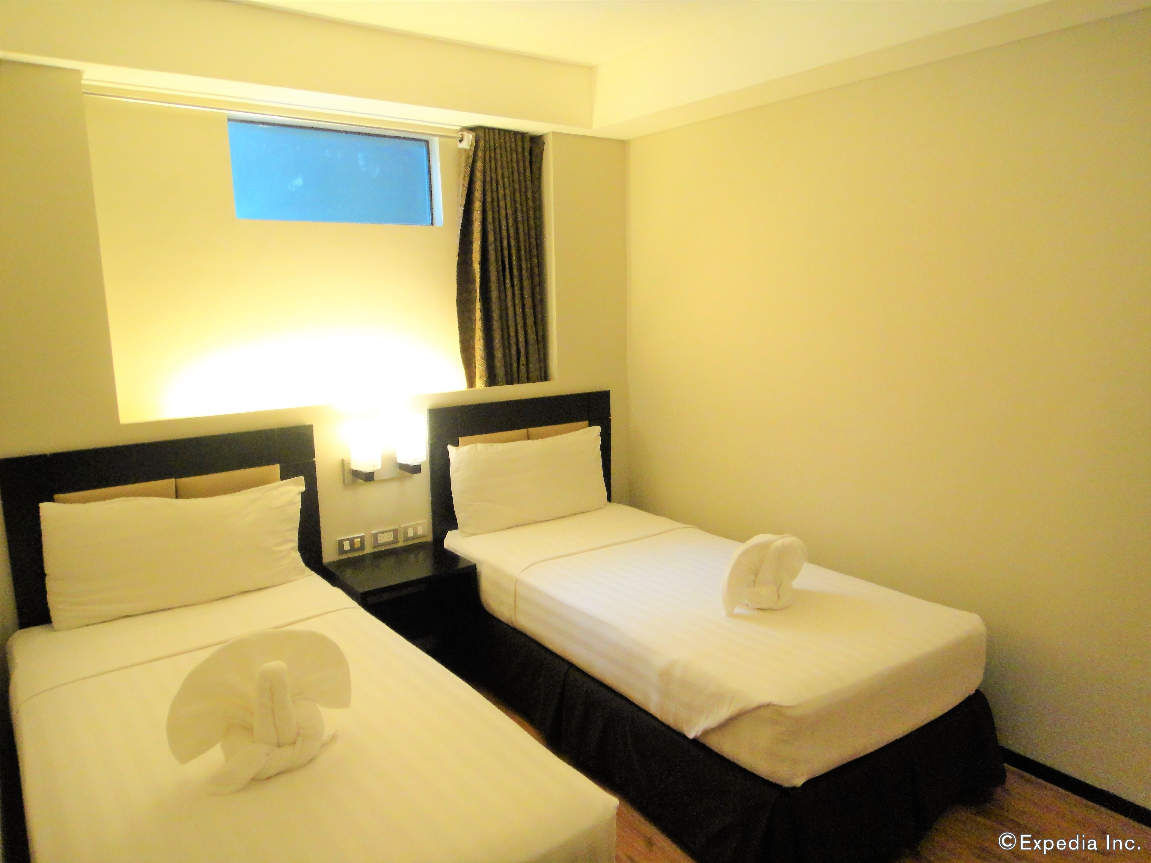 A hotel room with two single beds, a nightstand, a window with curtains, and a wall-mounted light.