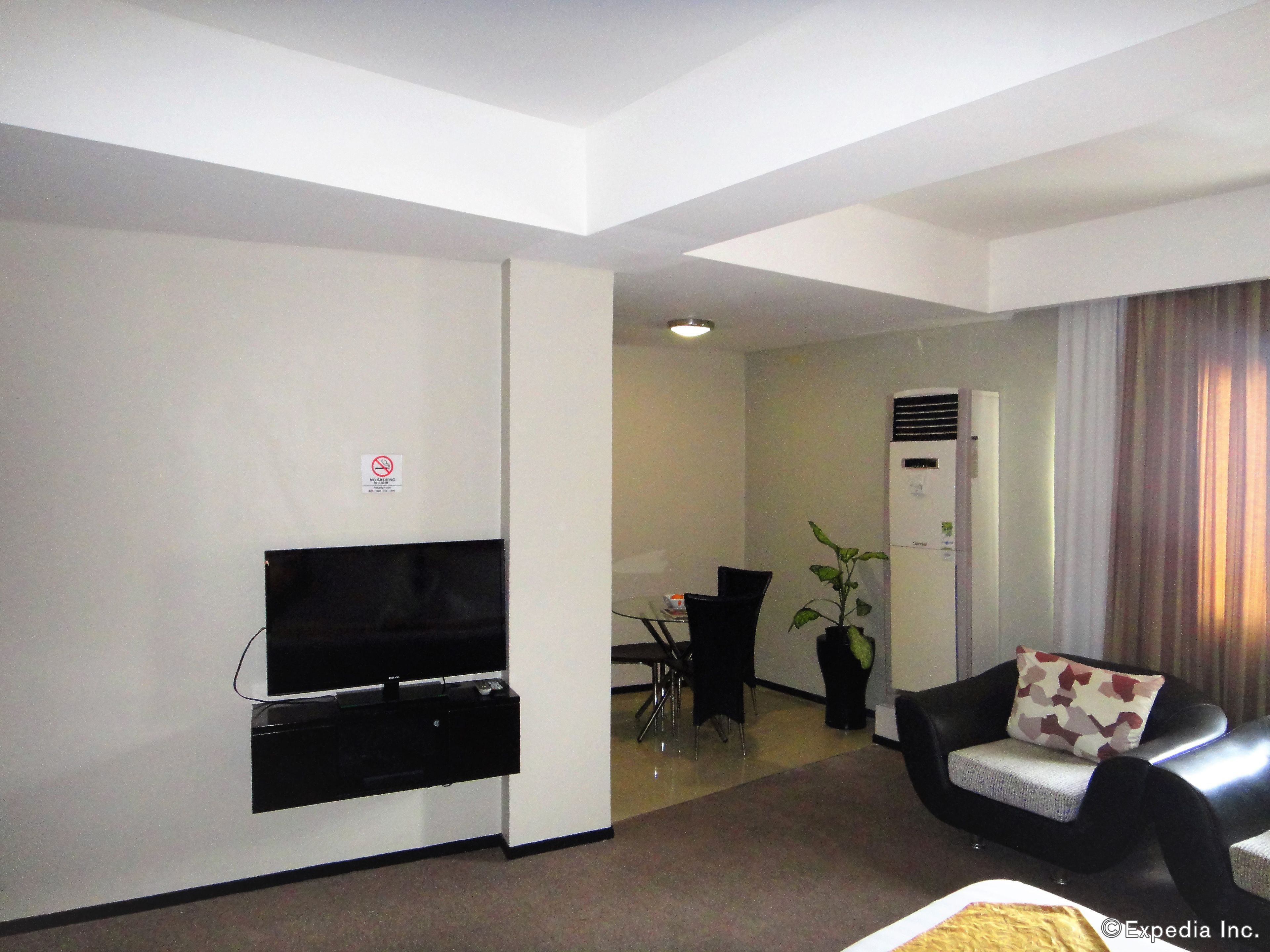 Un salon moderne avec un téléviseur à écran plat fixé au mur, un canapé noir agrémenté d’un coussin à motifs, une plante en pot et un coin repas avec une table et des chaises.