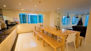 Una cocina moderna con una mesa de comedor de madera, bancos y una silla blanca. Hay ventanas con cortinas azules, un árbol de Navidad y un sofá negro en la sala.