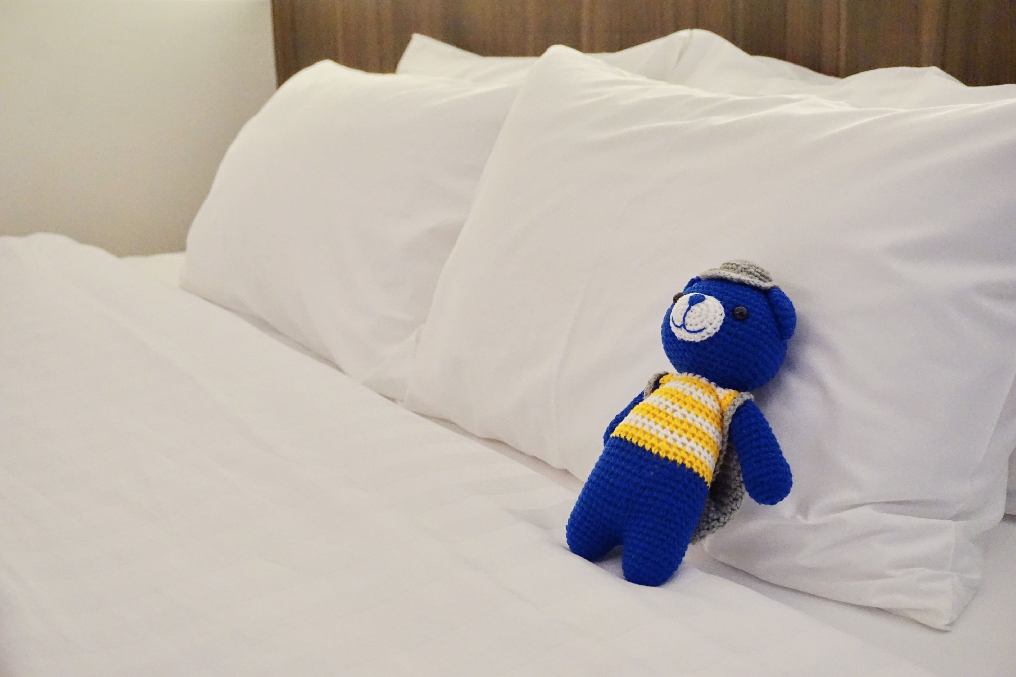 A blue teddy bear with a yellow and white striped shirt sitting on a white bed with pillows.