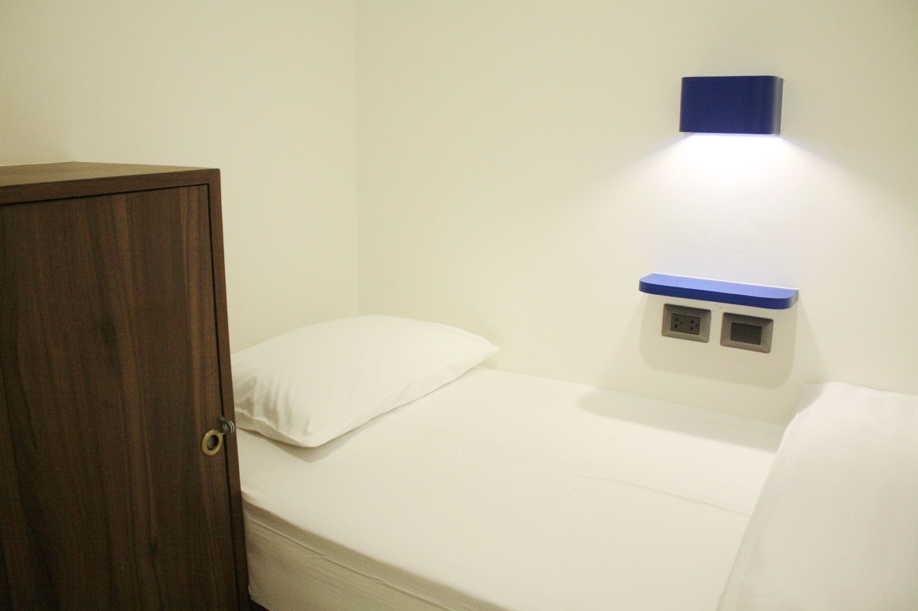 A small, single-bed room with a wooden wardrobe, a white pillow, and a wall-mounted electrical outlet.