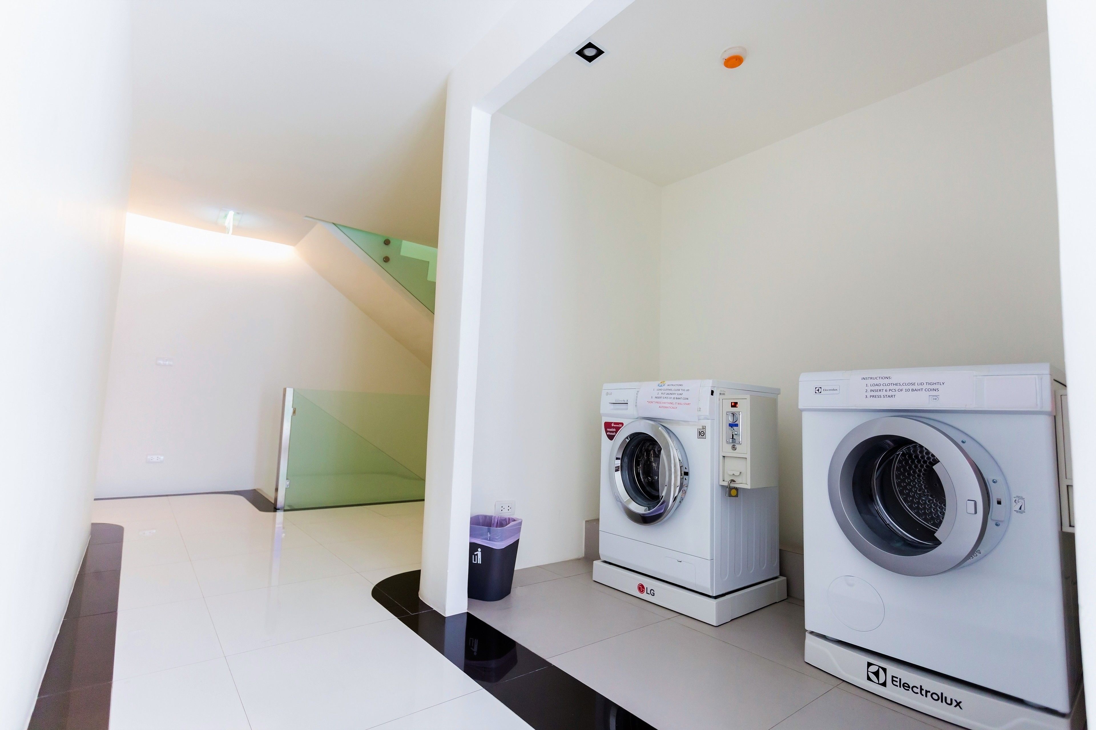 A laundry area with two washing machines, a trash bin, and a staircase.