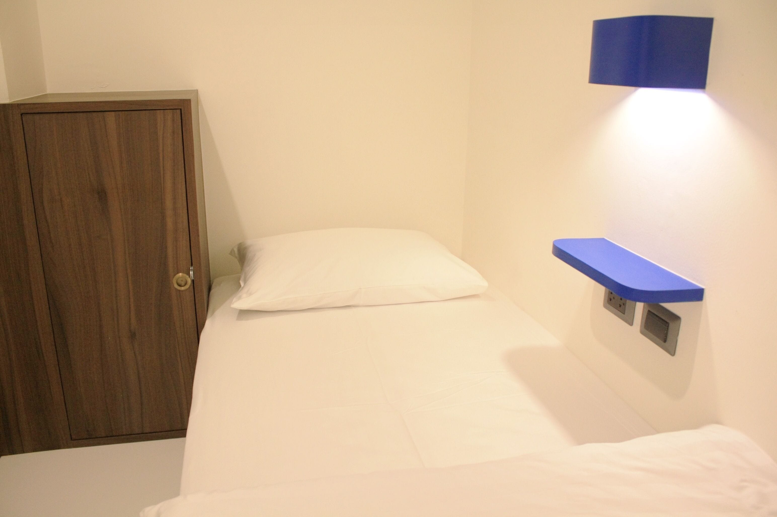 A small, neatly made bed with a white mattress and pillow, a wooden wardrobe, and a blue shelf with a light above it.