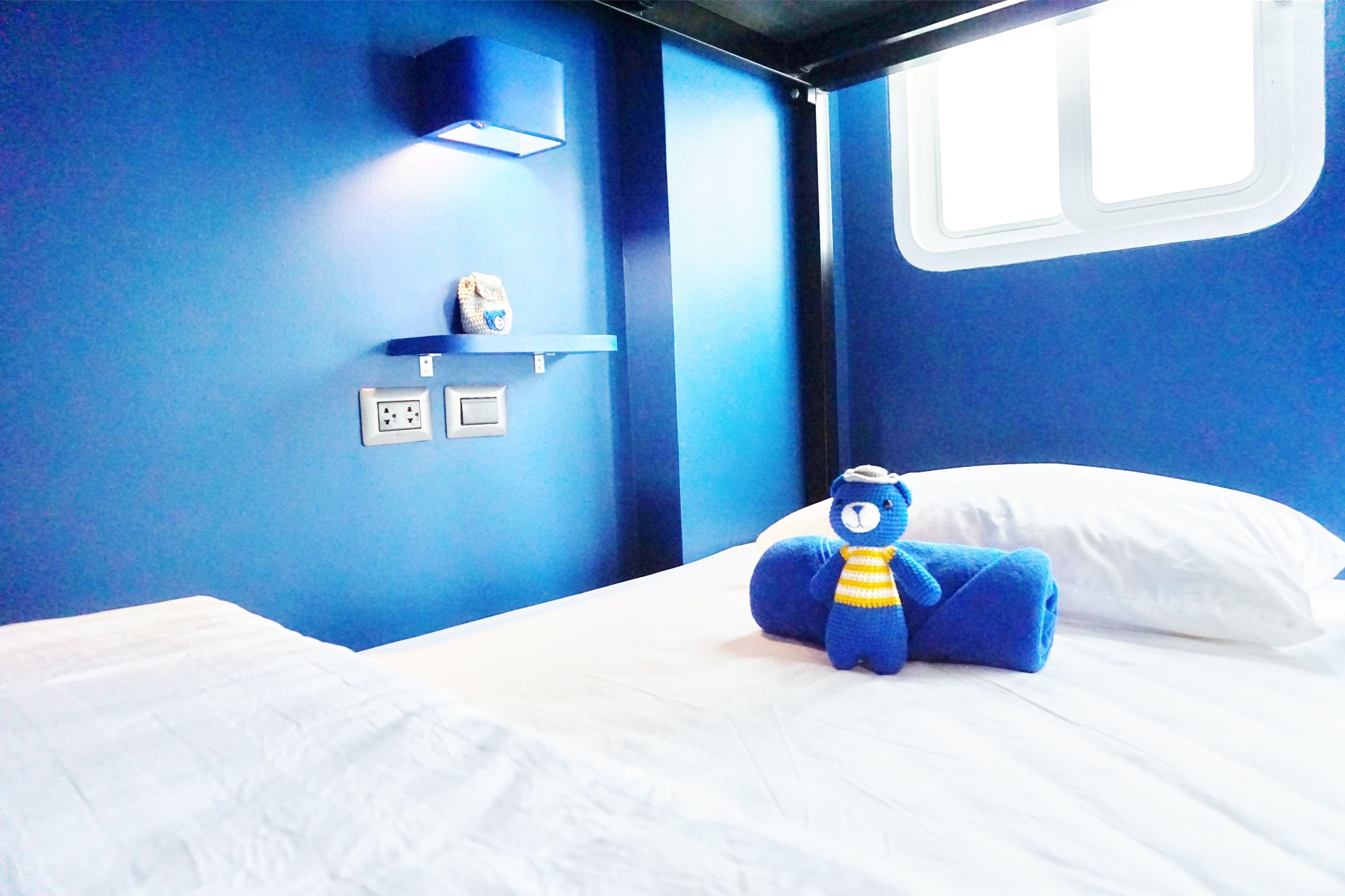 A bed with white bedding, a blue teddy bear, a shelf with a clock, and a window with blinds.