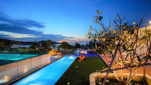 Una piscina con tobogán de agua, rodeada de jardines con una palmera y vistas de la ciudad al fondo.