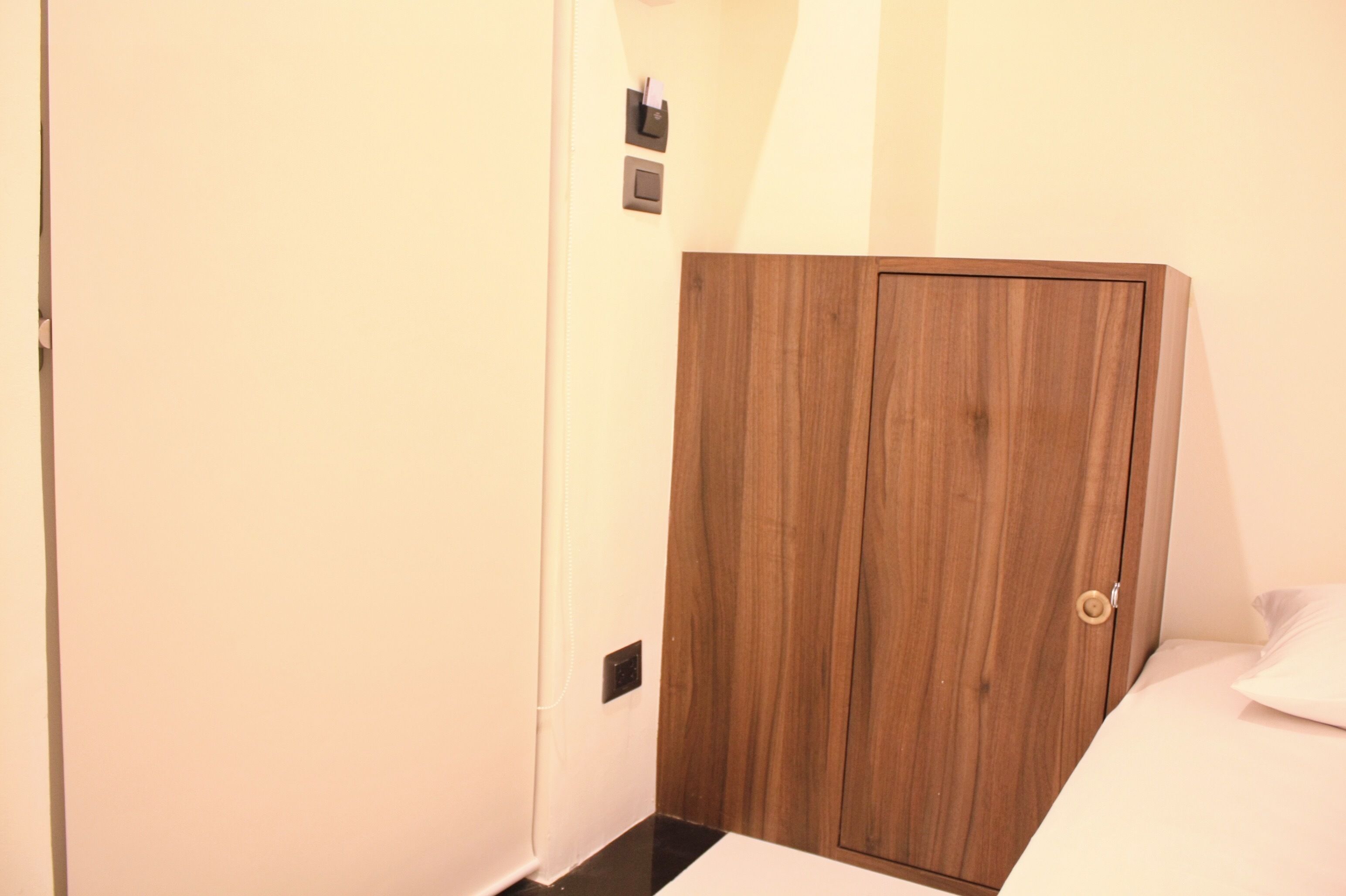 A room with a wooden wardrobe, a bed with white bedding, and a light switch on the wall.