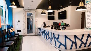 A reception area with a white counter, blue geometric patterns, and black pendant lighting. There are two blue chairs with orange accents and a small table with a laptop and a red box.