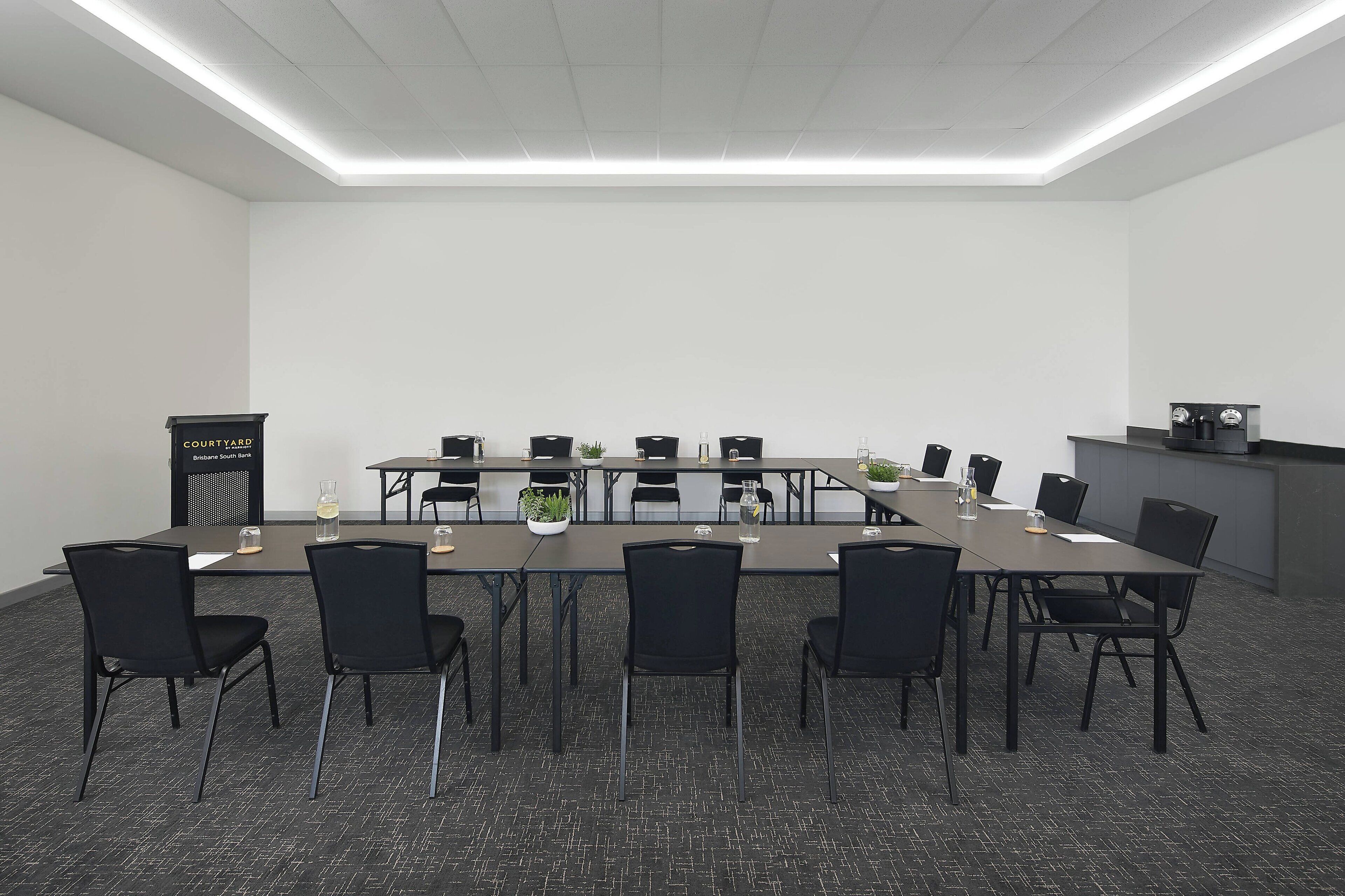 A conference room with a long table, black chairs, a podium, and a kitchenette area.