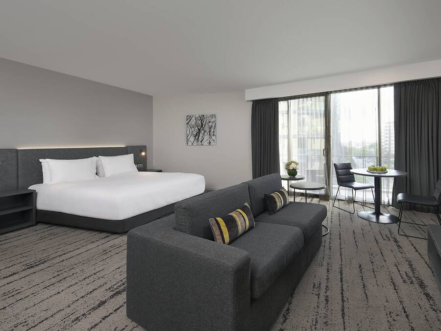 Una habitación de hotel moderna con una cama grande, un sofá, una mesita y vistas a la ciudad a través de amplios ventanales.