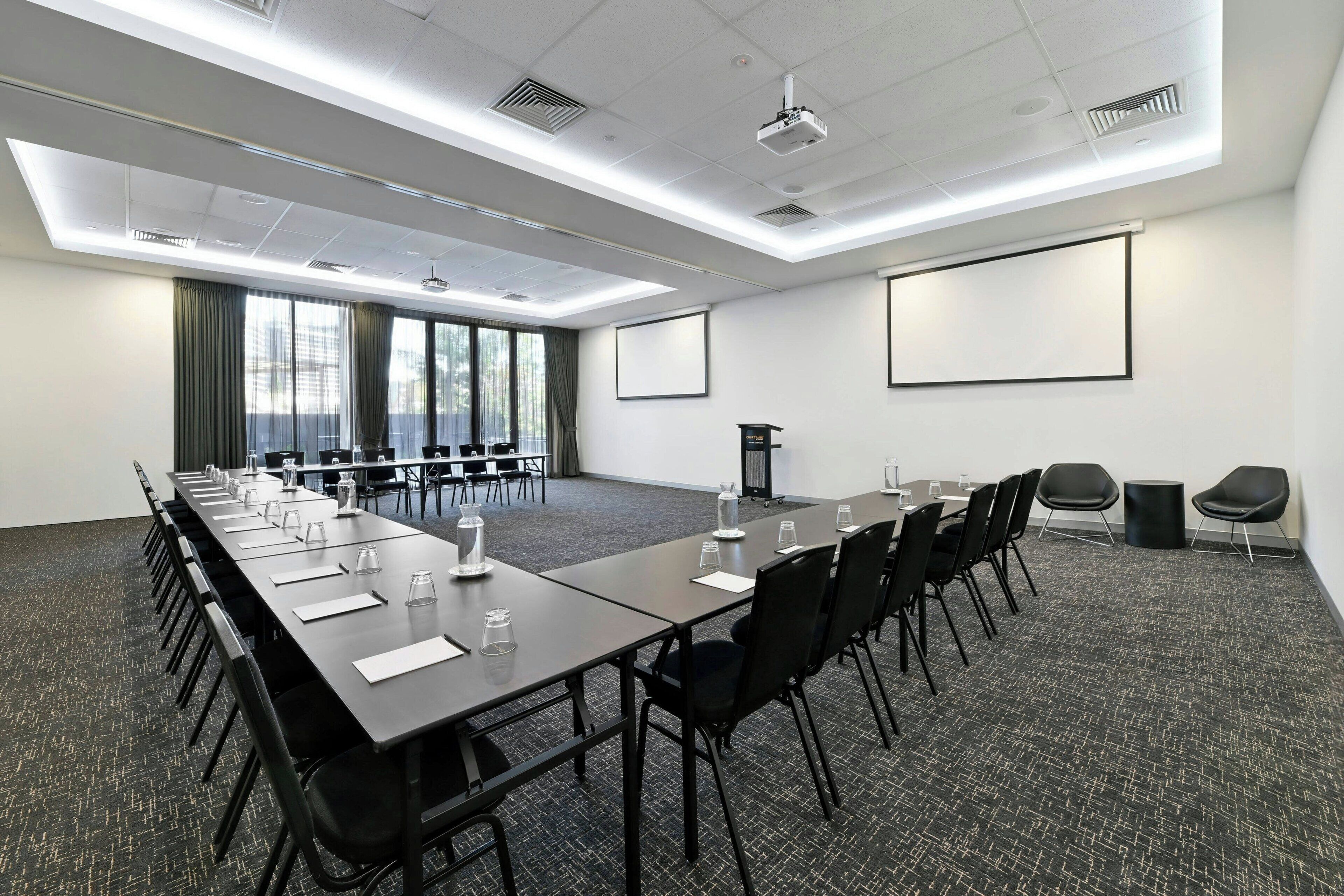 Un salón de conferencias con una mesa larga, sillas, una pantalla de proyector y un gran ventanal con cortinas.