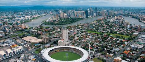 A stadium with a central field surrounded by a circular track, adjacent to a dense urban area with high-rise buildings and a river.
