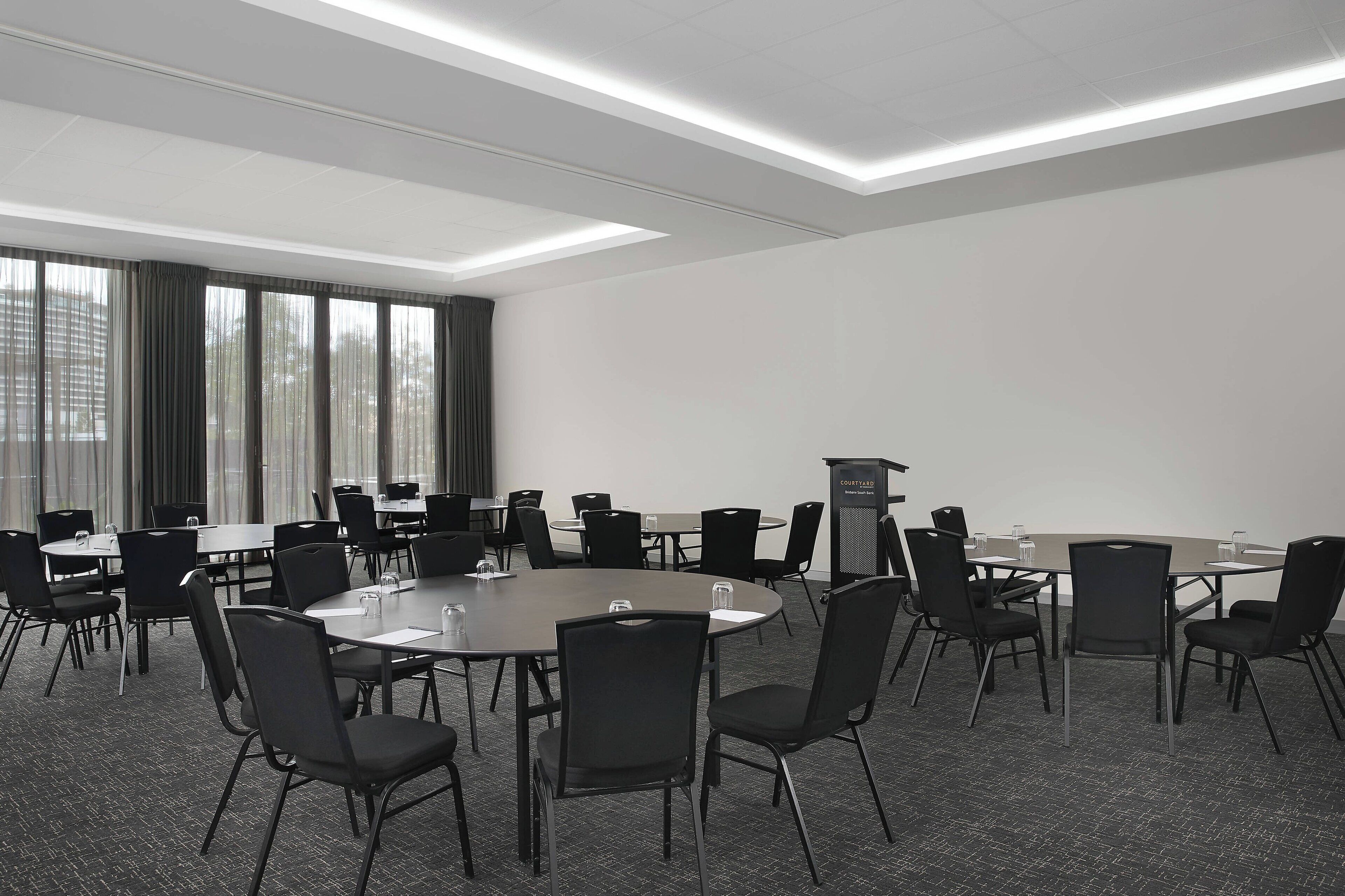 Un salón de conferencias con varias mesas y sillas, un atril y ventanas amplias con cortinas.
