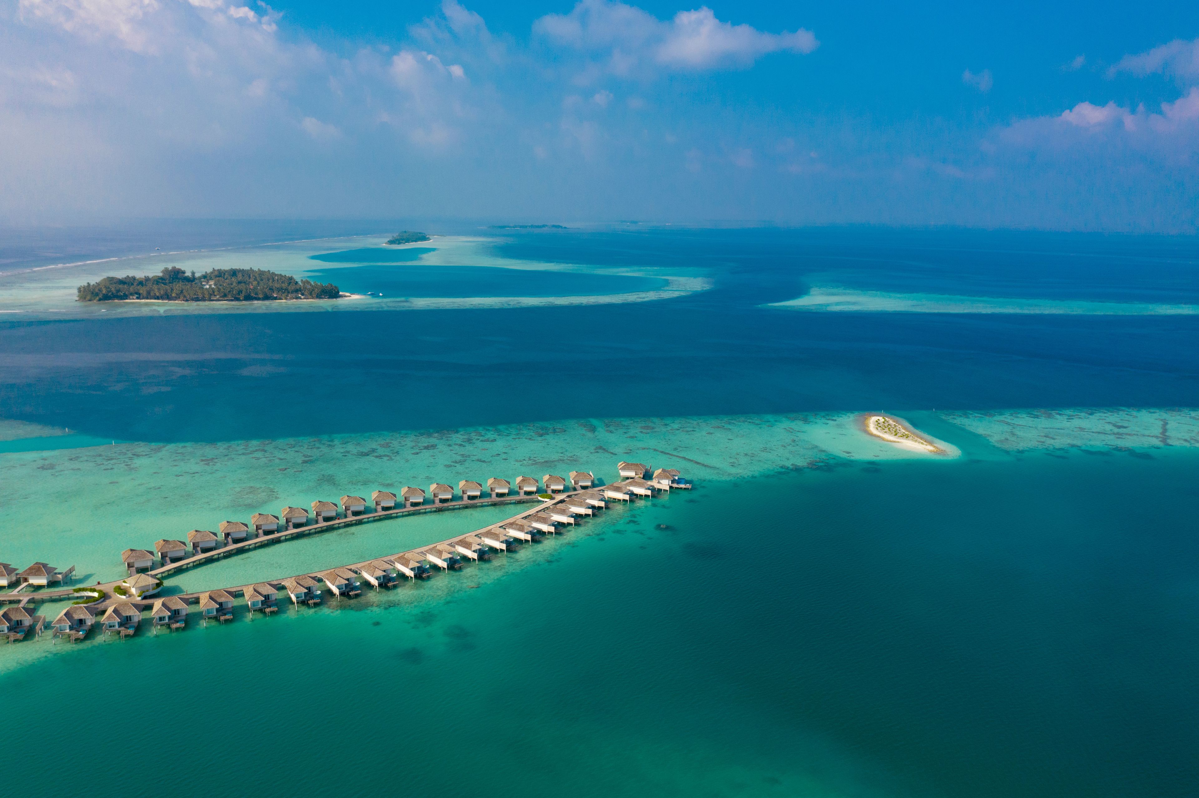 Vista aérea de un complejo turístico con bungalows sobre el agua conectados por un muelle, rodeado de aguas turquesas y pequeñas islas.