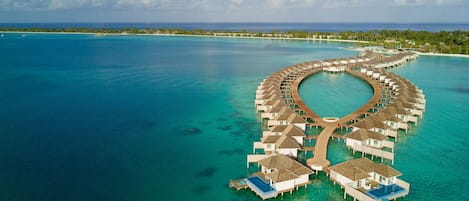 An overwater resort with a circular layout of bungalows connected by a wooden walkway.