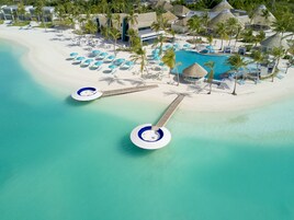 Vista aérea de un complejo turístico con una piscina grande, varios sombrillas en la playa y palmeras.