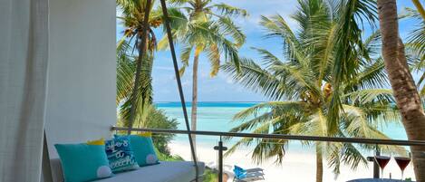 A balcony with a view of the beach, palm trees, and a lounge area with cushions and a table with drinks.